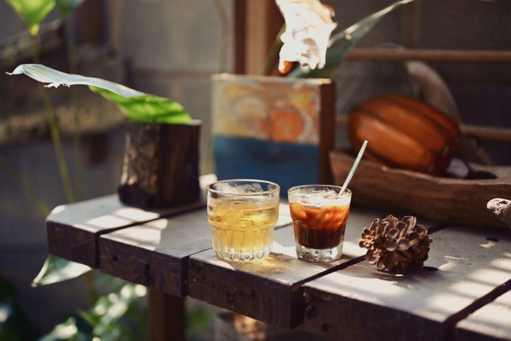 Close-up of cold beverages on a wooden table with tropical plants in a sunny outdoor setting.